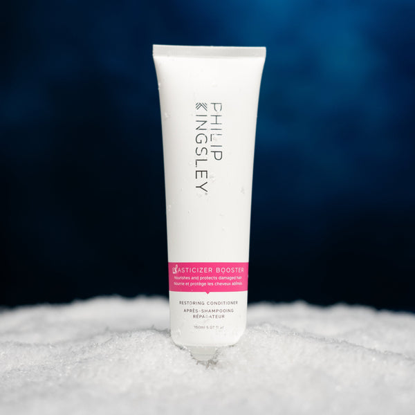 A white tube of Philip Kingsley Justicizer Boosting Restoring Conditioner standing upright on a textured white surface against a dark blue gradient background.