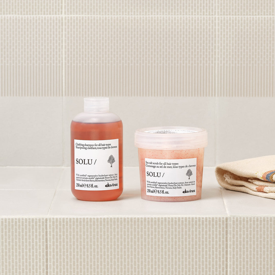Two Solu haircare products from the Solaire line are displayed on a beige tiled shelf next to a folded cream towel. The set includes a bottle of conditioning shampoo in translucent pinkish-orange plastic and a tub of hair growth serum in clear plastic with a white lid.