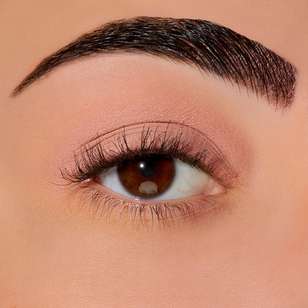 Extreme close-up of a human eye with a neatly shaped dark brown eyebrow, long eyelashes, and warm beige eyeshadow on a smooth skin surface.