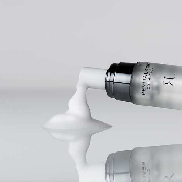Revitalash cosmetic bottle with white cream being squeezed out on a light gray background