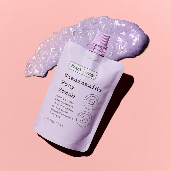 Purple packet of frank body Niacinamide Body Scrub with a dollop of scrub on a pink background