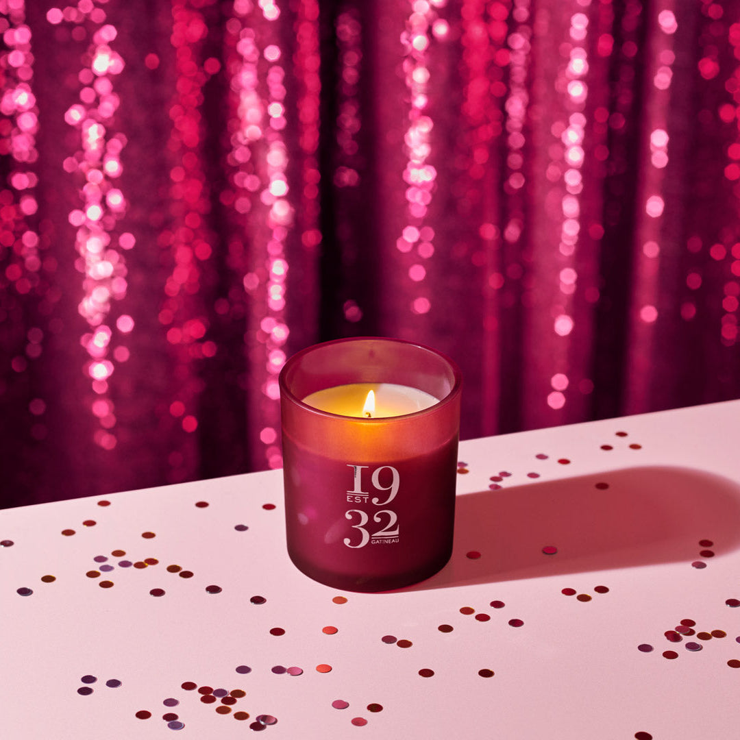Candle in a purple holder with '1932' on it, placed on a surface with confetti against a pink sequin curtain background.