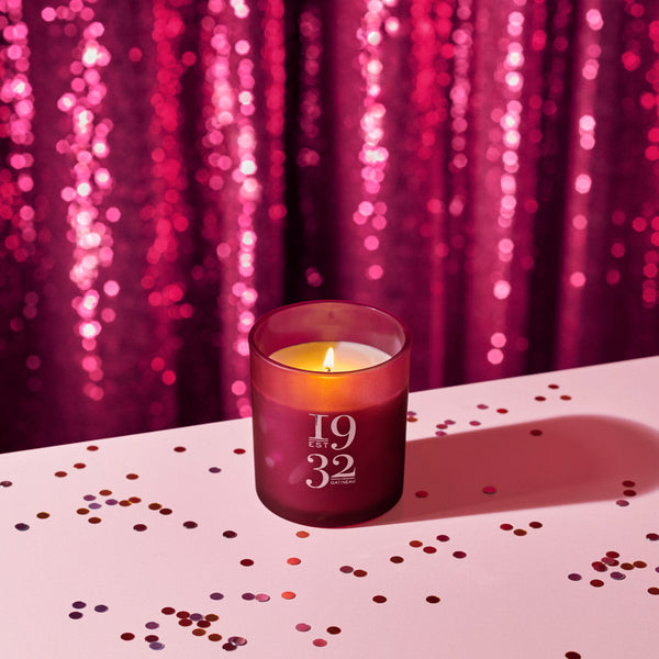 Candle in a purple holder with '1932' on it, placed on a surface with confetti against a pink sequin curtain background.