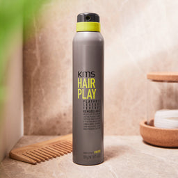 KMS Hair Play product bottle on a bathroom counter with a wooden comb and bowl in the background.