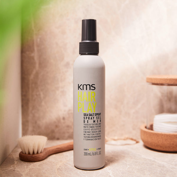 KMS Hair Play Sea Salt Spray bottle on a bathroom counter with a brush and soap dish in the background.