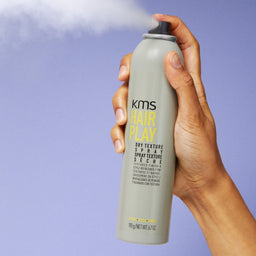 Hand holding a KMS Hair Play spray can against a purple background