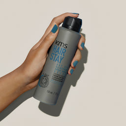 Hand holding a KMS Hair Stay product against a neutral background