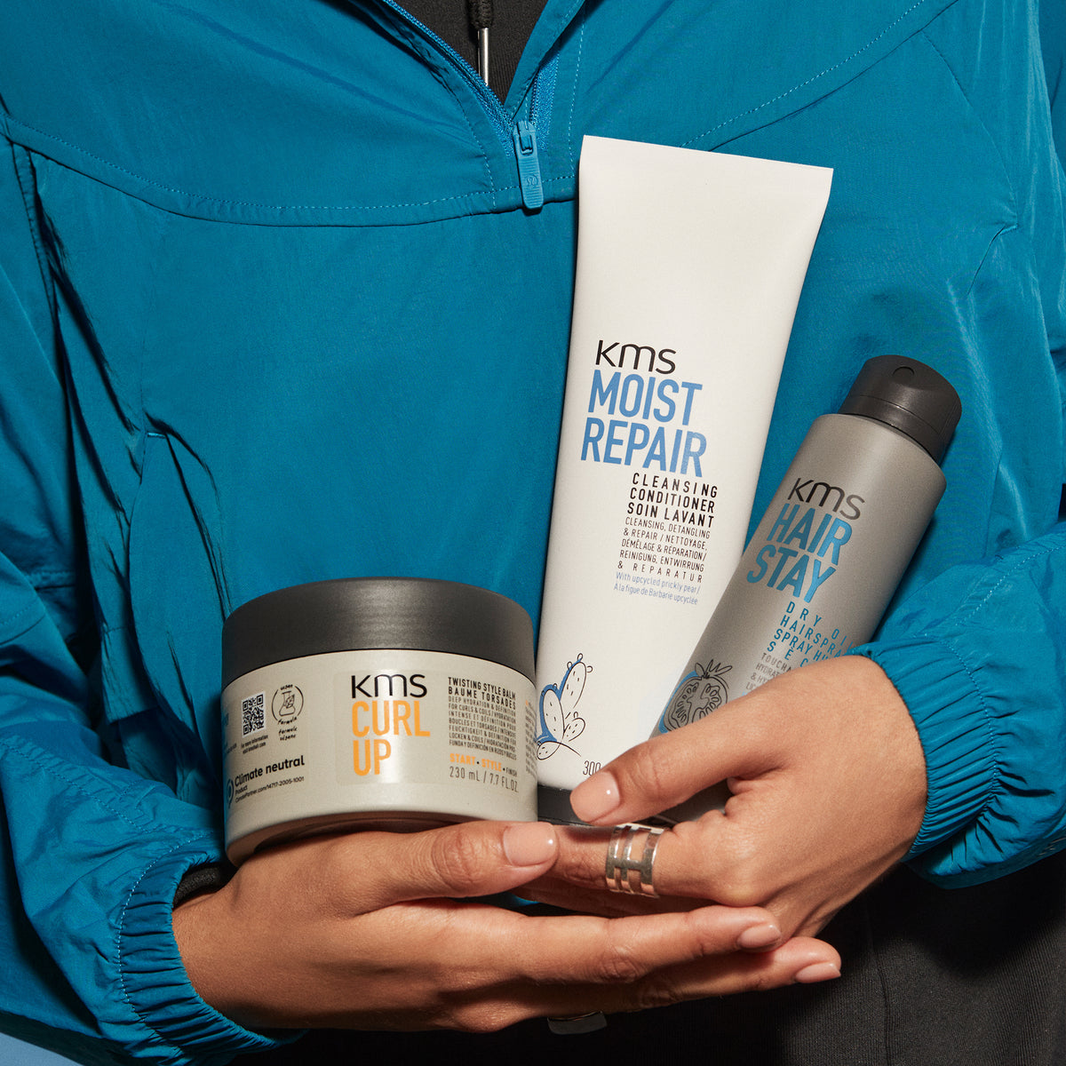 Person holding KMS hair care products including 'Moist Repair', 'Hair Stay', and 'Curly Up' in front of a blue jacket.