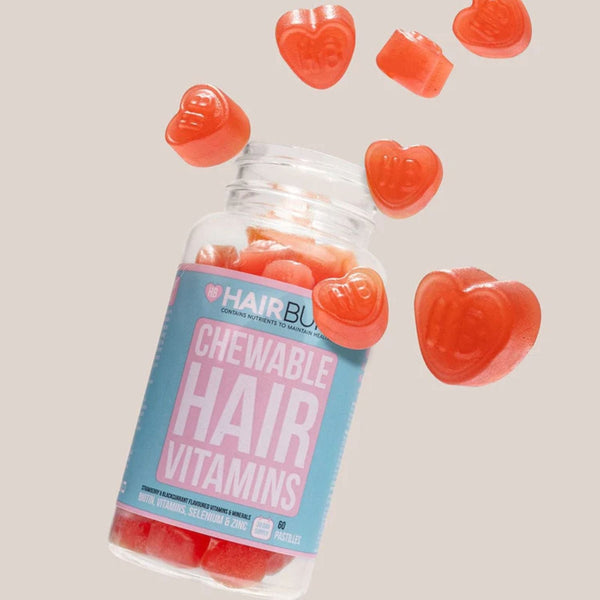 A clear bottle of HairBlast Chewable Hair Vitamins with a blue and pink label, shown with several red heart-shaped gummy vitamins floating around it on a light beige background.