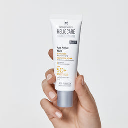A hand holding a white tube of Cantabria Bio Heliocare Age Active Fluid SPF 50+ sunscreen against a light gray background.