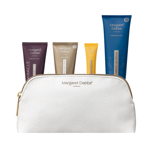 A white textured zippered cosmetic pouch with gold hardware, featuring the Margaret Dabbs London logo, with four colorful product tubes (purple, beige, yellow, and blue) arranged behind it on a white background.