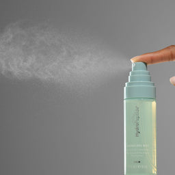 A hand pressing the spray nozzle of a Hydrophilic Hydration Mist bottle, with a fine mist spraying out against a solid gray background.