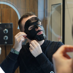 Man applying a black face mask in front of a mirror.