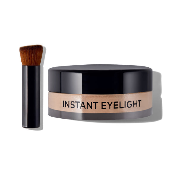 Instant Eyelight makeup jar with a black lid and a beige band label, shown next to a small black makeup brush with brown bristles on a white background.