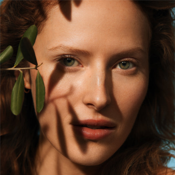 Close-up of a woman's face with a leaf in front of her eye, sunlit.