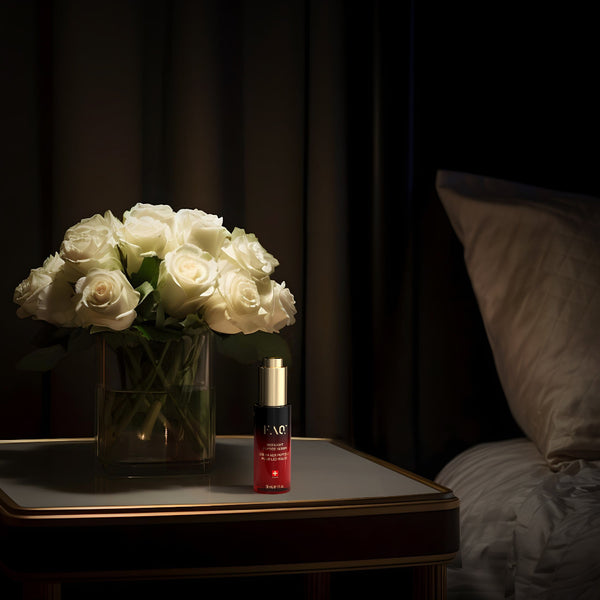 A small red cosmetic bottle with a gold cap stands on a bedside table next to a vase of white roses, with a dimly lit bed and pillow in the background.