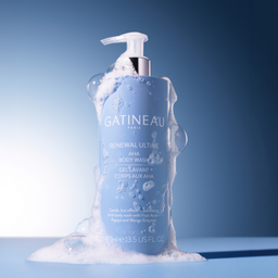 A bottle of Gatineau Renewal Ultime AHA Body Wash, filled with light blue liquid and covered in white foam, set against a blue gradient background.
