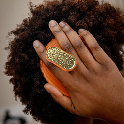 A close-up lifestyle shot of a person with dark curly hair wearing a large, chunky gold ring with an orange band. The ring features a textured, hammered oval face with a polished gold center.
