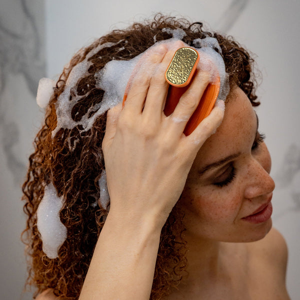 A woman with curly brown hair lathering it with white foam while using an orange and gold handheld scalp brush against a light marble-tiled wall.