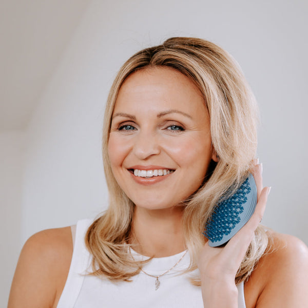 A smiling blonde woman holds a blue silicone hair massager against her hair, wearing a white sleeveless top against a bright, neutral indoor background.