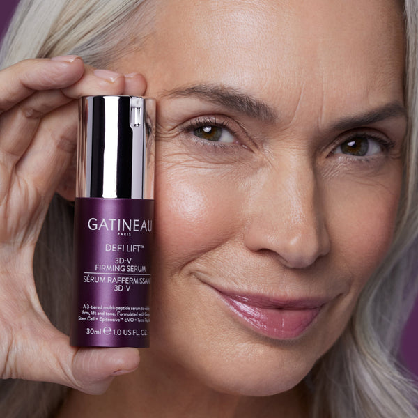 A close-up lifestyle shot of a woman holding a bottle of Gatineau Paris Defi Lift 3D-V Firming Serum next to her face.