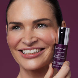 A close-up of a smiling woman holding a bottle of Gatineau Defi Lift 3D-Y Revitalizing Serum against a solid dark purple background.