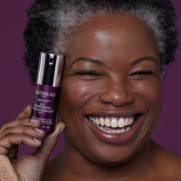 A smiling woman with short gray hair holds a bottle of GATINEAU Deep Lift 3D-V Cellular Serum against a solid purple background.