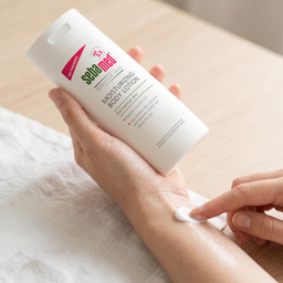 A person holds a white bottle of Seta Med Moisturising Body Lotion over their forearm, with a dollop of the white lotion being spread onto the skin.