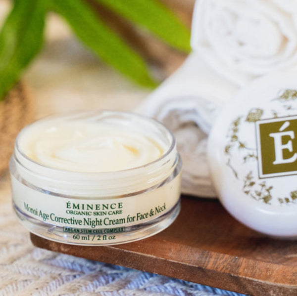 A jar of Eminence Organic Skin Care Manicure Age Corrective Night Cream next to its white lid, which features a green monogram logo, displayed on a wooden tray with rolled white towels and green leaves in the background.