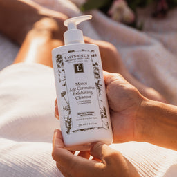 A person holds a white pump bottle of Eminence Organic Body Care Monoi Age Corrective Exfoliating Cleanser against a warm, sunlit background of skin and soft fabric.