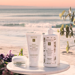 Three Eminence Organic Beauty Care products arranged on a white round table by the ocean: a small jar, a tube labeled Moroi Age Corrective Night Body Cream, and a pump bottle labeled Moroi Age Corrective Exfoliating Cleanser, with beach waves and flowers in the background.