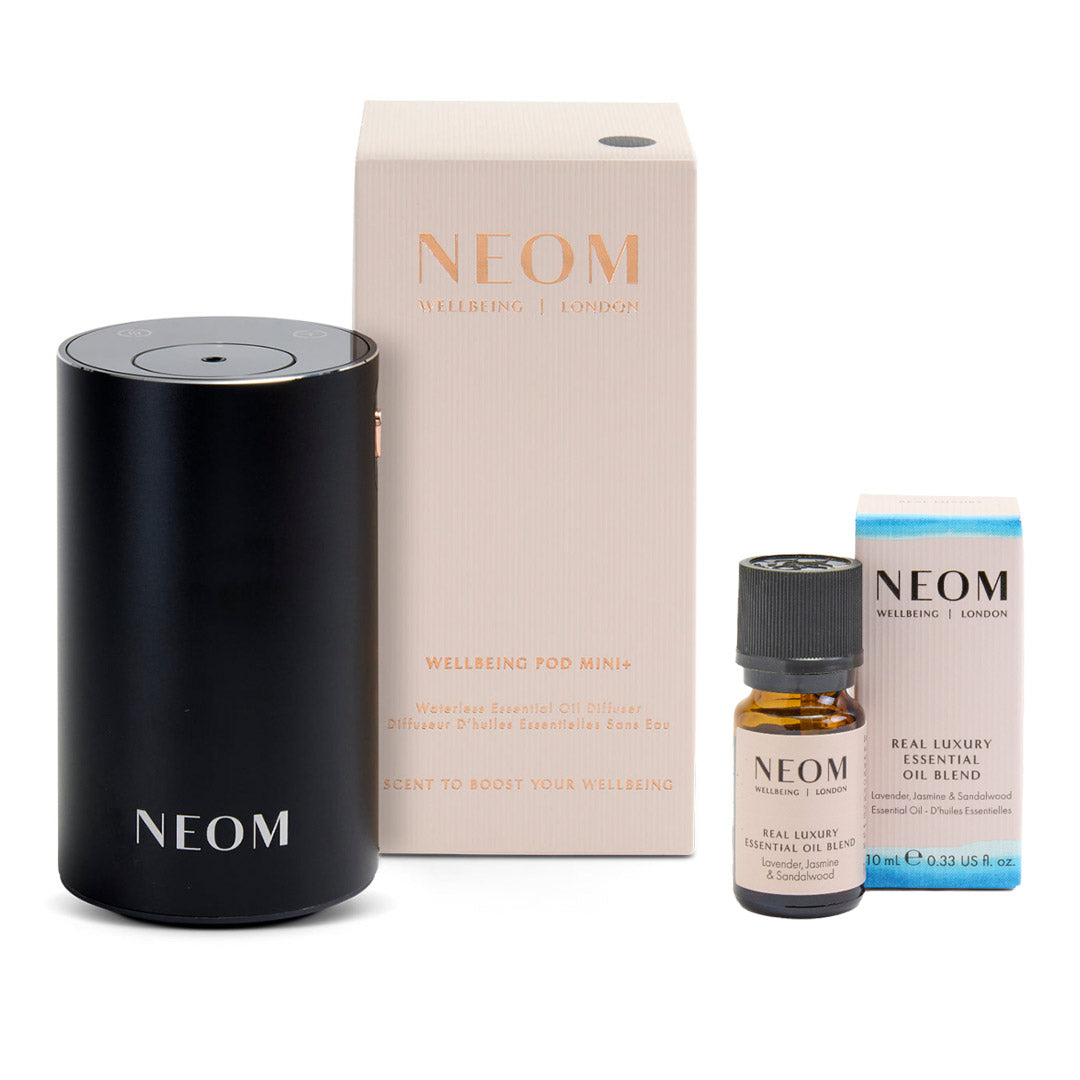 NEOM Welcoming Pod Mini diffuser set on a white background, including the matte black cylindrical diffuser, its cream-colored packaging box, a small amber essential oil bottle, and a blue-accented product box.