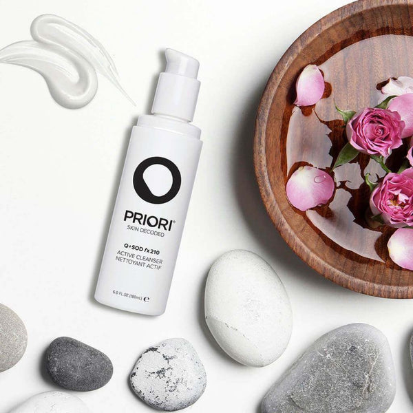 a bottle of PRIORI Q+SOD - Active Cleanser next to a bowl of flower petals and stones