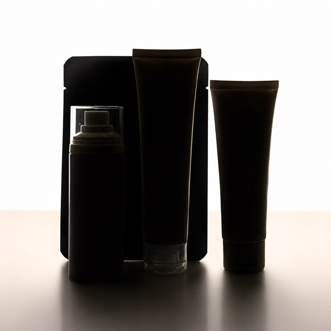 Three dark cosmetic or toiletry containers standing against a bright white background: a rectangular pouch, a tall squeeze tube, and a cylindrical bottle with a clear cap.