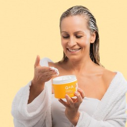 Woman in a white robe applying cream from a yellow container with 'Q+A' branding on a yellow background