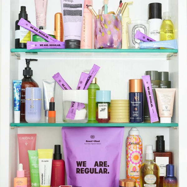 A white shelving unit filled with assorted skincare, cosmetics, and beauty products, including tubes, bottles, brushes, and a large purple bag in front with the text 'WE ARE. REGULAR.