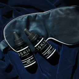 two OSKIA Retinoid Sleep Serum Level 1 - 0.2% bottles on a blue silk cloth and blue eyemask
