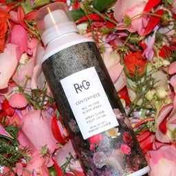 a bottle of R+Co Centerpiece All-In-One Hair Elixir on top of a bed of petals