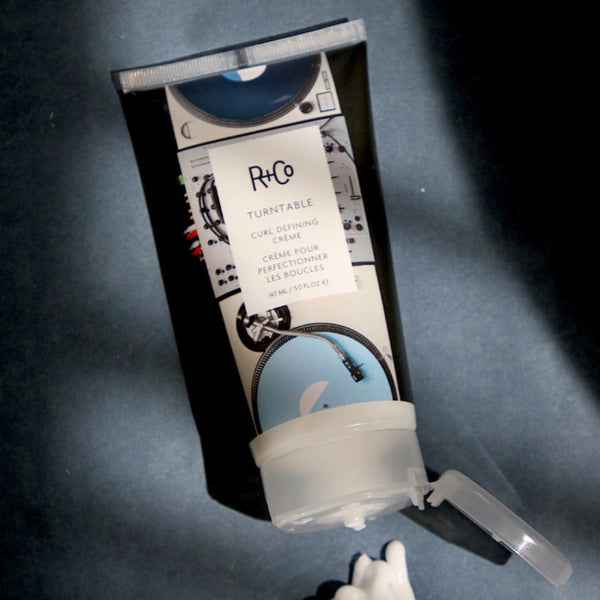 R+Co Turn Table Curl Defining Cream tube on a dark blue surface