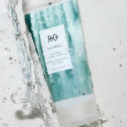 a tube of R+Co Waterfall Moisture + Shine Lotion under a stream of water