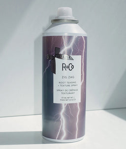 a can of R+Co Zig Zag Root Teasing + Texture Spray in the corner of a white surface 