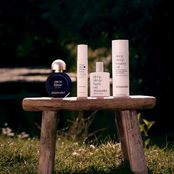 Four skincare products from thisworks are arranged on a rustic wooden bench outdoors on grass with a dark, blurred natural background. The products include a round dark blue bottle with a gold cap labeled 'own time', a white cylindrical bottle, a white rectangular bath oil bottle, and a tall white beauty oil bottle.