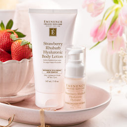 Eminence Organic Skin Care Strawberry Rhubarb Hyaluronic Body Lotion and matching serum bottle displayed on a pink tray with strawberries and pink flowers in the background.