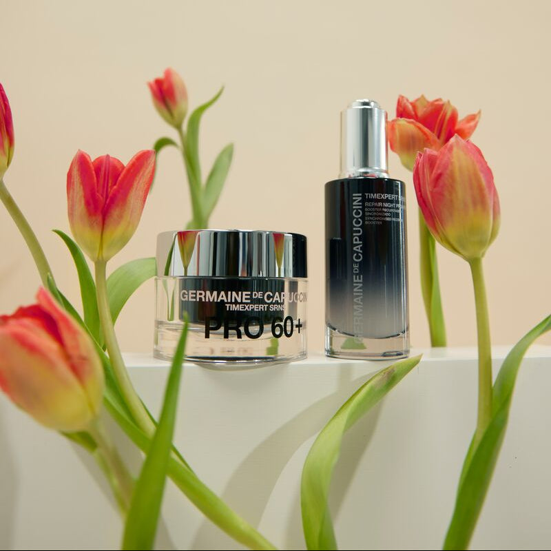 Two skincare products on a white surface with red tulips in the foreground.