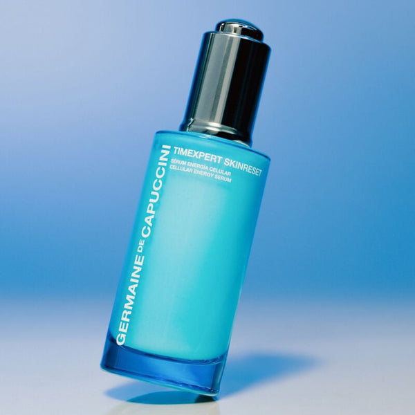 A bottle of Germaine de Capuccini Timexpert Skinreset serum, featuring a translucent light blue glass bottle and a reflective silver pump dispenser, set against a clean blue gradient background.