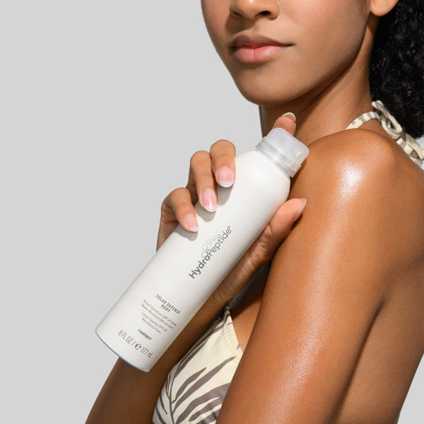 Woman applying sunscreen spray to her arm with a neutral background