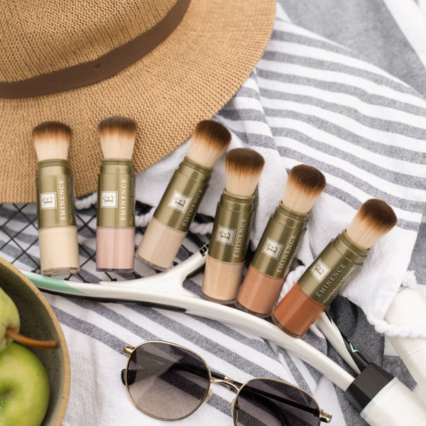 Six makeup brushes with olive green handles and brown bristles are arranged on a white and gray striped towel, alongside a straw hat, sunglasses, and a bowl of green apples.