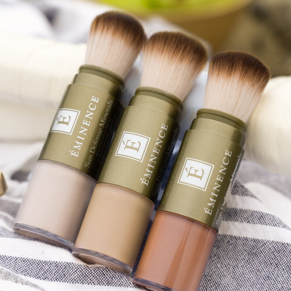 Three Eminence makeup brushes with sponge-tip bristles in beige, tan, and medium brown shades, displayed on a striped fabric background with a blurred bowl of green apples in the background.
