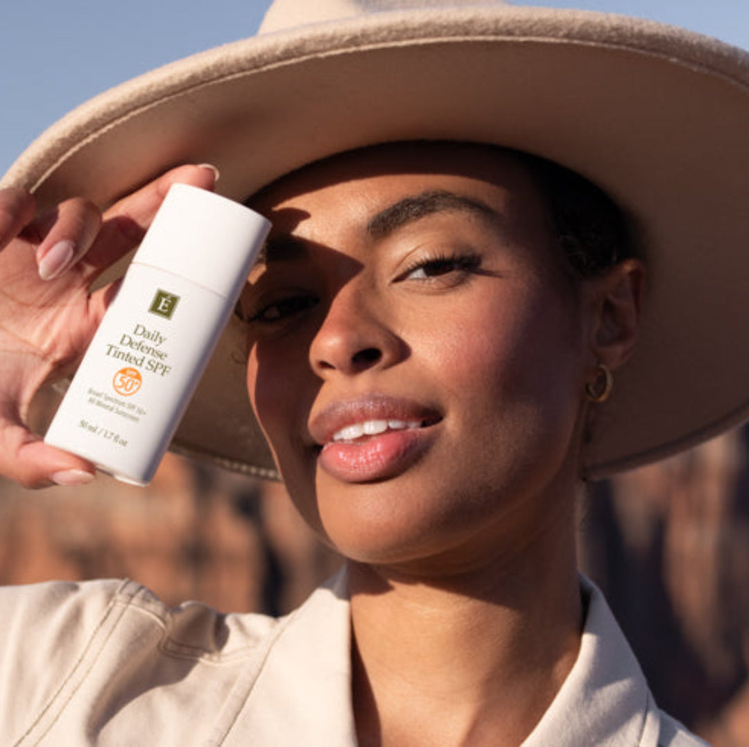 A woman wearing a beige wide-brim hat and matching top holds a white bottle of JUVENIAL Daily Sun Protection SPF 50+ in front of her face, with a desert canyon landscape in the background.