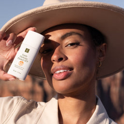 A woman wearing a beige wide-brim hat and matching top holds a white bottle of JUVENIAL Daily Sun Protection SPF 50+ in front of her face, with a desert canyon landscape in the background.
